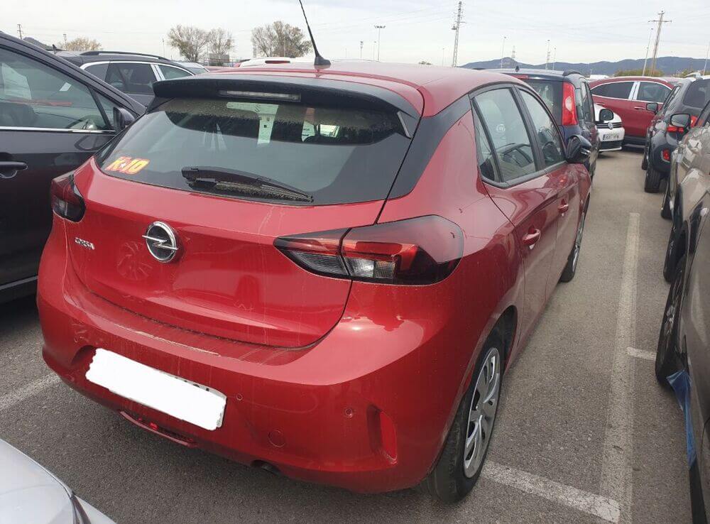 Una imagen de la parte trasera de un Opel seminuevo estacionado en una calle de Zaragoza. El coche es de color rojo y muestra un diseño moderno con líneas aerodinámicas. Se pueden ver claramente las luces traseras y el logotipo de Opel. En el fondo, hay una vista urbana típica de Zaragoza con edificios y árboles alineados en la calle. La imagen destaca la condición bien cuidada del vehículo.
