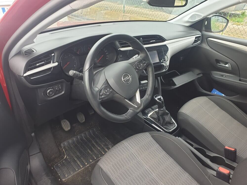 Una imagen del interior de un Opel Corsa de segunda mano. La imagen muestra los asientos delanteros de color gris oscuro y el tablero de mandos con un diseño moderno y funcional. El volante tiene controles integrados, y la consola central incluye una pantalla táctil para el sistema de entretenimiento y navegación. Los materiales del interior parecen estar en buen estado, reflejando el cuidado del coche.