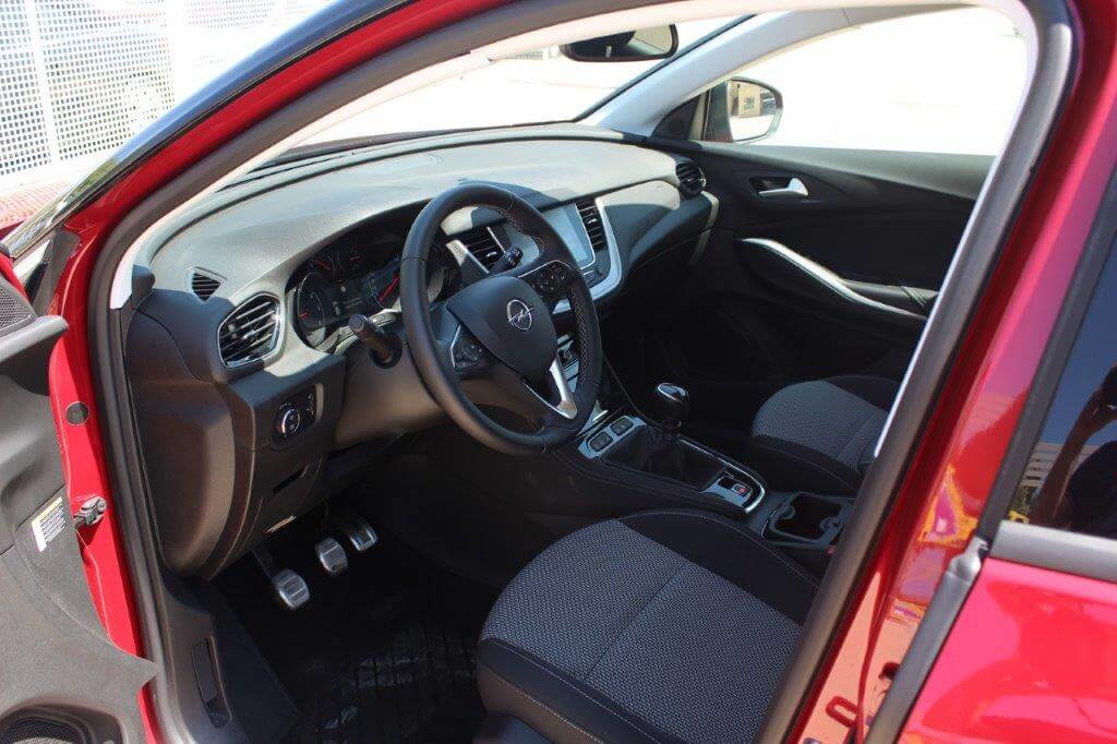 El interior de un Opel Grandland X 2021 seminuevo, con la puerta del conductor abierta. La imagen muestra el volante, el tablero de instrumentos, la consola central y los asientos delanteros. El volante tiene el logotipo de Opel en el centro y varios controles integrados. La consola central incluye una palanca de cambios manual, portavasos y controles adicionales. Los asientos son de tela con un diseño de dos tonos. La imagen es relevante porque proporciona una vista detallada del interior del vehículo, destacando su diseño y características.