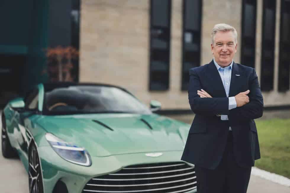 La imagen muestra a Adrian Hallmark, el CEO de Aston Martin, de pie frente a un coche verde Aston Martin. Hallmark está vestido con un traje oscuro y una camisa a cuadros de color azul claro. Sus brazos están cruzados y tiene una expresión seria. Al fondo, se puede ver un edificio con grandes ventanas, que parece ser parte de las instalaciones de Aston Martin.