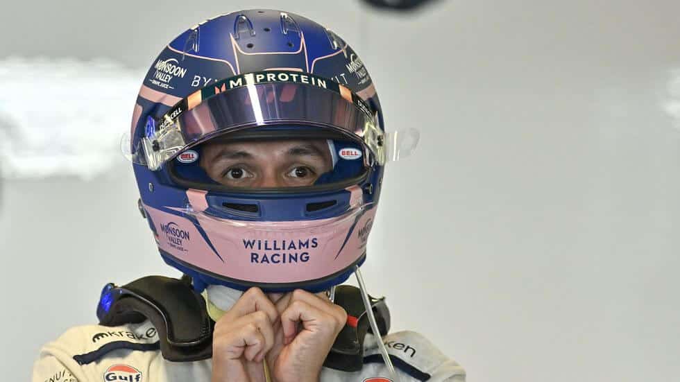 Alex Albon, piloto de carreras, ajustándose el casco antes de una competición. El casco es azul con detalles en rosa y tiene varios patrocinadores visibles, como 'My Protein' y 'Monsoon Valley'. Albon lleva un traje de carreras blanco con logotipos de patrocinadores como 'Gulf' y 'mikra'. La imagen captura un momento de preparación crucial antes de una carrera de automovilismo.