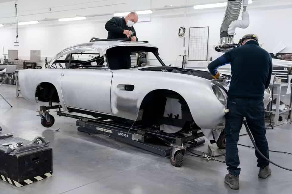 La imagen muestra a dos personas trabajando en la restauración o ensamblaje de un coche clásico Aston Martin en un taller. El coche está montado en una plataforma móvil, y las personas están realizando tareas que parecen involucrar la carrocería y los detalles. Una persona trabaja en el techo del coche, mientras que la otra está enfocada en la parte delantera. El taller está bien iluminado y equipado con diversas herramientas y maquinaria, lo que indica un entorno profesional para la restauración automotriz.