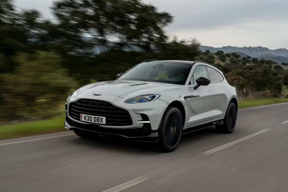 "La imagen muestra un Aston Martin DBX blanco conduciendo por una carretera con un fondo borroso de árboles y montañas, lo que indica movimiento. La matrícula del coche dice 'K30 DBX'. La imagen destaca el modelo SUV de lujo de Aston Martin, resaltando su diseño elegante y capacidades de rendimiento.