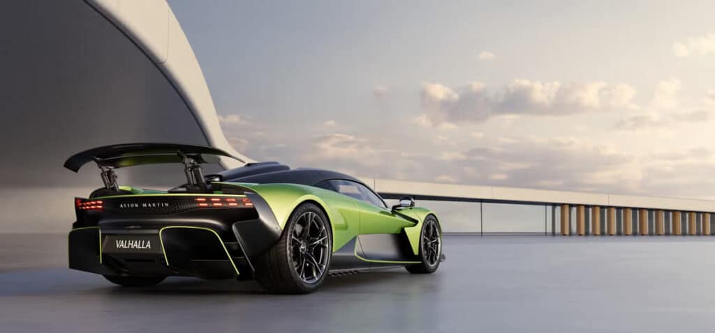 "La imagen muestra un automóvil deportivo Aston Martin Valhalla de color verde y negro, visto desde un ángulo trasero-lateral. El coche está estacionado en una superficie amplia y lisa, con una estructura arquitectónica moderna al fondo. Se destacan el diseño aerodinámico del automóvil, el alerón trasero prominente y las distintivas luces traseras. La matrícula dice 'VALHALLA' y el nombre de la marca 'ASTON MARTIN' es visible encima de las luces traseras. El ambiente parece ser al atardecer o al amanecer, con un cielo despejado y una iluminación suave, lo que añade un toque dramático y lujoso a la imagen.