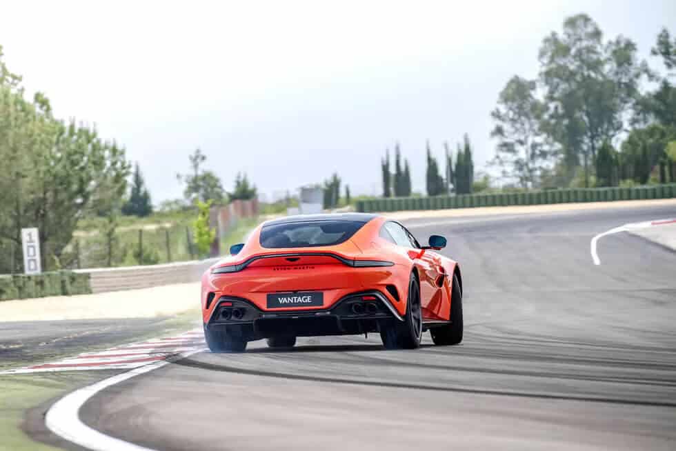 "La imagen muestra un Aston Martin Vantage de color naranja, conduciendo en una pista de carreras. El coche se ve desde atrás mientras toma una curva, con un ligero derrape indicado por el humo que sale de los neumáticos traseros. La pista está rodeada de vegetación y árboles, con un cartel en el lado izquierdo que muestra el número '100'. La imagen resalta el rendimiento y la capacidad de manejo del Aston Martin Vantage en un escenario de conducción dinámica.