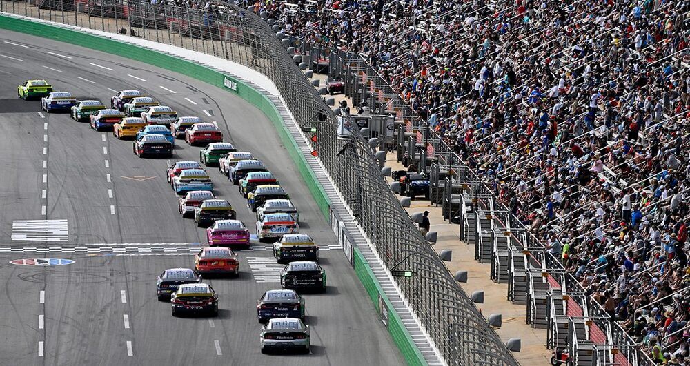 Imagen de una carrera de NASCAR en progreso. Varios coches de carreras están en la pista, todos moviéndose en la misma dirección y muy juntos, lo que indica una carrera competitiva. La pista está rodeada por una gran multitud de espectadores sentados en las gradas a la derecha de la imagen. La pista tiene varios carriles y marcas en el asfalto, incluyendo una línea de inicio/meta. La imagen captura la emoción e intensidad de una carrera de NASCAR, destacando la gran cantidad de participantes y la audiencia entusiasta.