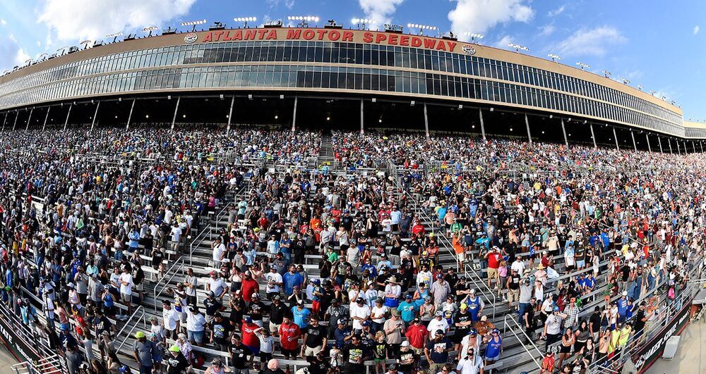 Imagen de una multitud de espectadores en el Atlanta Motor Speedway. Las gradas están llenas de personas, indicando un evento significativo, probablemente una carrera de NASCAR. El cartel del Atlanta Motor Speedway se muestra de manera prominente en la parte superior de las gradas. La imagen captura la escala y la emoción del evento, destacando la popularidad y la gran asistencia a las carreras de NASCAR.