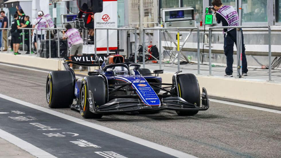 El coche de Fórmula 1 del equipo Williams Racing en el pit lane de un circuito de carreras. El coche es principalmente azul con varios logotipos de patrocinadores y el número 23 en el morro. Se pueden ver varios miembros del equipo y fotógrafos en el fondo, lo que indica que probablemente sea durante una sesión de práctica, clasificación o una carrera. El pit lane está marcado con varias líneas y símbolos, y hay un cartel con el logotipo de MoneyGram en el fondo. Esta imagen es interesante y relevante ya que captura un momento de un evento de automovilismo de alto perfil, mostrando la tecnología avanzada y el trabajo en equipo involucrado en la Fórmula 1.