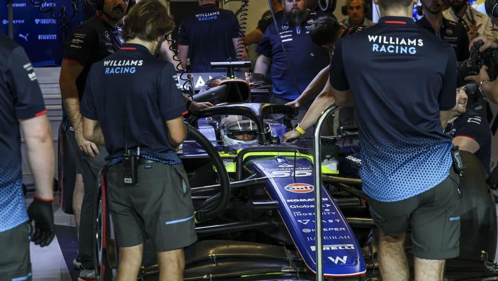 "Un grupo de miembros del equipo Williams Racing trabajando en un coche de Fórmula 1 en el garaje. Los miembros del equipo llevan uniformes con la inscripción 'WILLIAMS RACING' en la espalda. El coche está siendo preparado o ajustado, con varios miembros del equipo atendiendo diferentes partes del vehículo. Esta imagen es interesante y relevante ya que captura un momento detrás de escena en el entorno de alta presión de la Fórmula 1, destacando el trabajo en equipo y la precisión necesarios en el automovilismo.