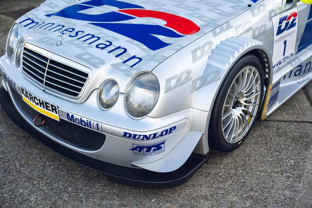 "Fotografía de un coche de carreras Mercedes CLK DTM de color plateado. La carrocería del vehículo está decorada con logotipos de patrocinadores como 'D2', 'mannesmann', 'Mobil 1', 'DUNLOP', 'ATS' y 'KÄRCHER'. El diseño aerodinámico del coche incluye un parachoques delantero prominente, faros redondeados y una parrilla con el emblema de Mercedes-Benz en el centro. La imagen está tomada desde un ángulo bajo, destacando el lateral derecho y la parte delantera del coche.