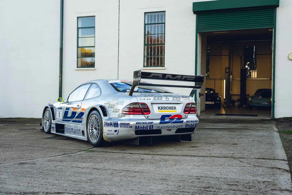 Fotografía de un coche de carreras Mercedes CLK DTM plateado, estacionado frente a un edificio con una puerta de garaje verde abierta. El auto tiene un diseño aerodinámico con un gran alerón trasero y está decorado con logotipos de patrocinadores, como 'Kärcher', 'Mobil 1', 'Dunlop' y 'Mannesmann'.