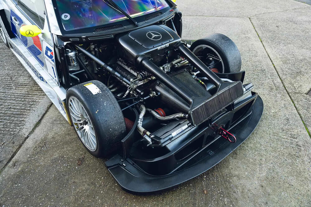 Vista detallada del Mercedes CLK DTM con el capó abierto, mostrando su motor y componentes mecánicos internos. El motor lleva el logotipo de Mercedes-Benz, rodeado de cables, tubos y un radiador en la parte frontal. Las llantas de aleación y neumáticos de carreras, notablemente desgastados, son visibles. La carrocería aerodinámica luce colores y logotipos de patrocinadores, destacando su diseño de competición.