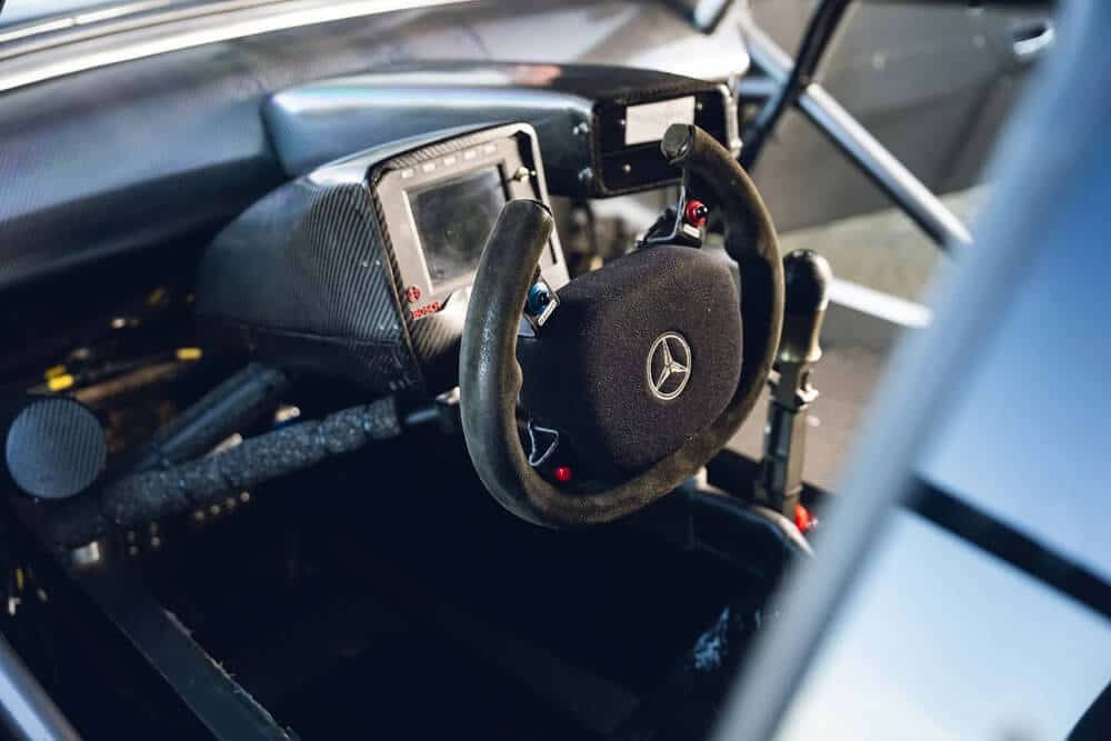 Interior de un coche de carreras Mercedes CLK DTM, centrado en el volante con el logotipo de Mercedes-Benz en el centro. Se puede ver un panel de instrumentos digital detrás del volante, junto con varios controles y botones alrededor del mismo, diseñados para ajustes en competición.