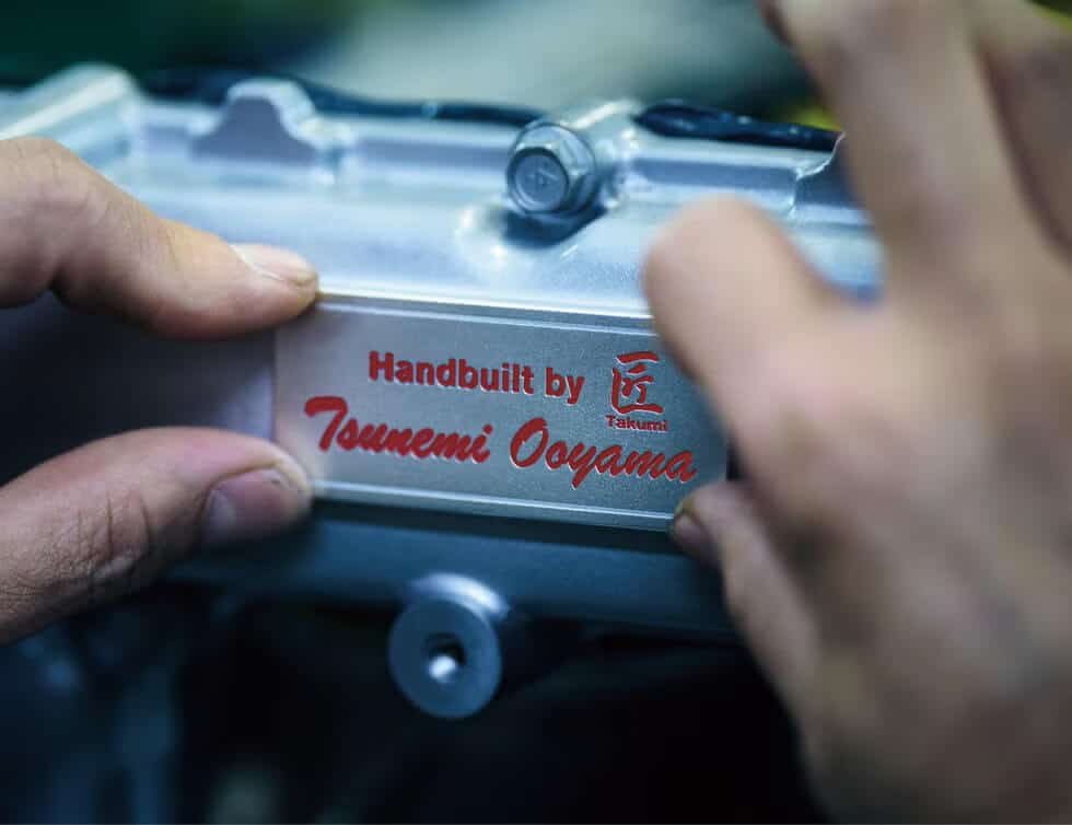 Primer plano del motor de un Nissan GT-R R35, destacando una placa metálica con la inscripción 'Handbuilt by Tsunemi Ooyama' y el logotipo de Takumi grabado. Dos manos sostienen la placa, simbolizando el trabajo artesanal y la dedicación detrás de este vehículo icónico, especialmente relevante en el contexto de su fin de producción.