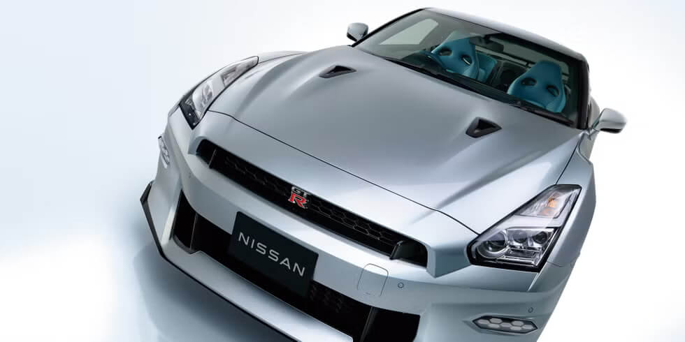 "Imagen de un Nissan GTR R35 plateado visto desde un ángulo frontal. El auto presenta un diseño deportivo y aerodinámico, con faros delanteros estilizados y una parrilla prominente que lleva el emblema 'GT-R' en el centro. El capó incluye dos ventilaciones que realzan su estilo dinámico, y a través del parabrisas se perciben asientos delanteros en color azul. Este modelo icónico se muestra en un contexto que marca el fin de su producción.