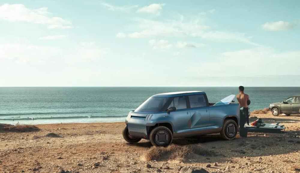 "Camioneta eléctrica azul de la marca 'Telo Trucks' estacionada en una playa con vistas al océano. En el fondo, se observa a varias personas disfrutando del agua. A la derecha de la camioneta, un hombre sin camisa está de pie junto a una tabla de surf y otros equipos colocados en el suelo. Otra camioneta está estacionada más lejos hacia la derecha, completando una escena costera tranquila y soleada.