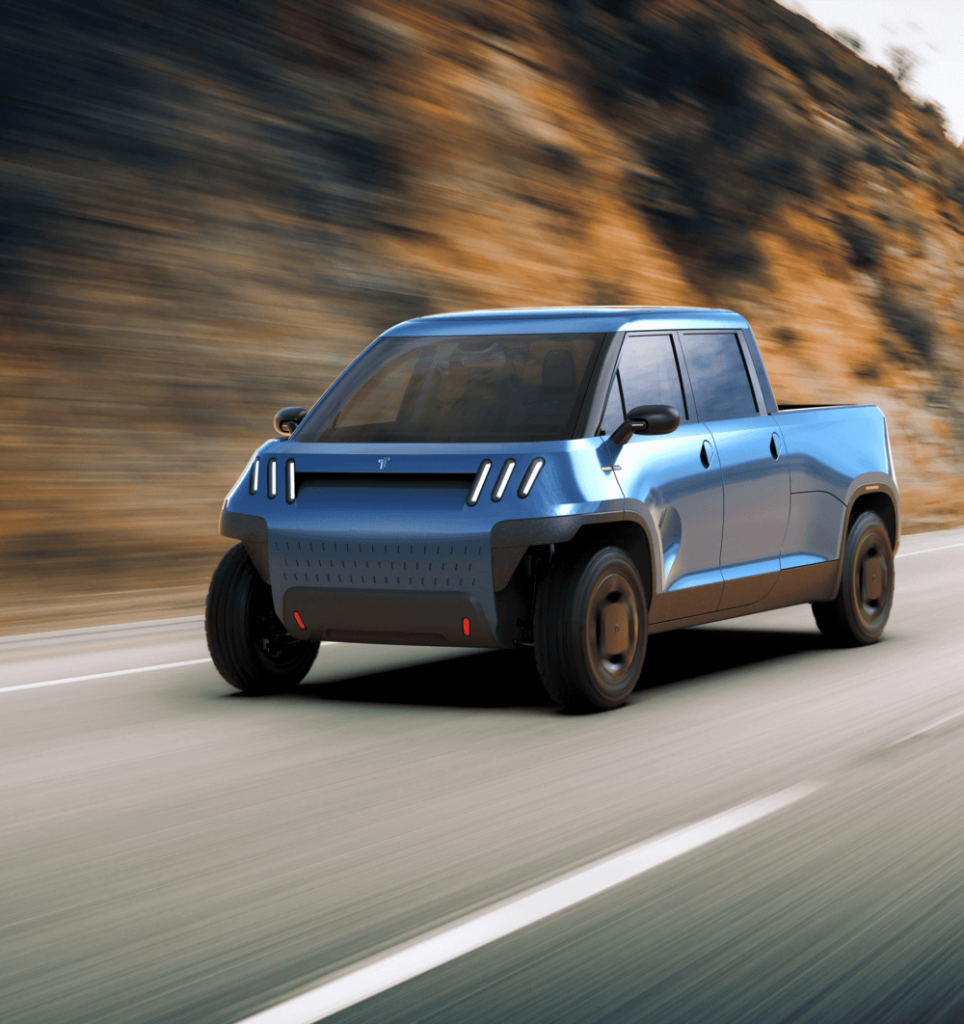 Camioneta eléctrica azul de la marca 'Telo Trucks' circulando por una carretera rodeada de un paisaje montañoso. El diseño moderno y aerodinámico del vehículo se destaca, con un frontal que incluye luces LED verticales y un parabrisas amplio que proporciona una vista clara del interior.
