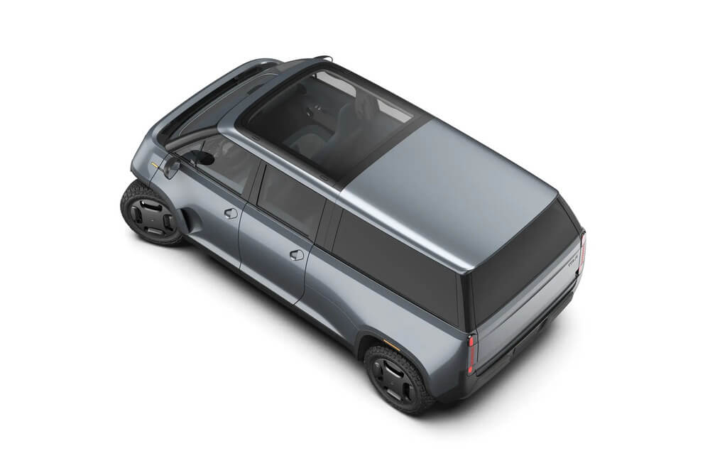 La imagen muestra una camioneta eléctrica llamada 'Telo Trucks' desde una vista aérea. La camioneta tiene un diseño elegante y moderno con una carrocería de color gris metálico. Destaca su techo parcialmente transparente y sus ventanas amplias, así como unas ruedas con un diseño distintivo y futurista.