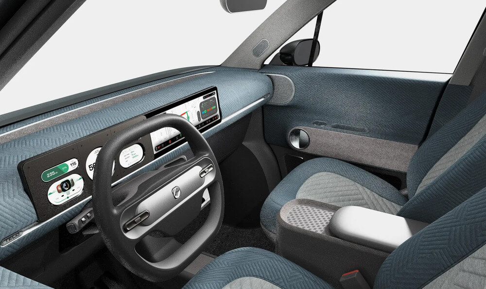 Vista interior de la camioneta eléctrica 'Telo Trucks', con un diseño moderno y minimalista. El salpicadero cuenta con una amplia pantalla digital que muestra información como velocidad, nivel de batería y otros indicadores del vehículo. El volante tiene un diseño elegante con controles integrados. Los asientos y superficies interiores están tapizados con un tejido texturizado en tonos azul y gris, resaltando una estética limpia y futurista.