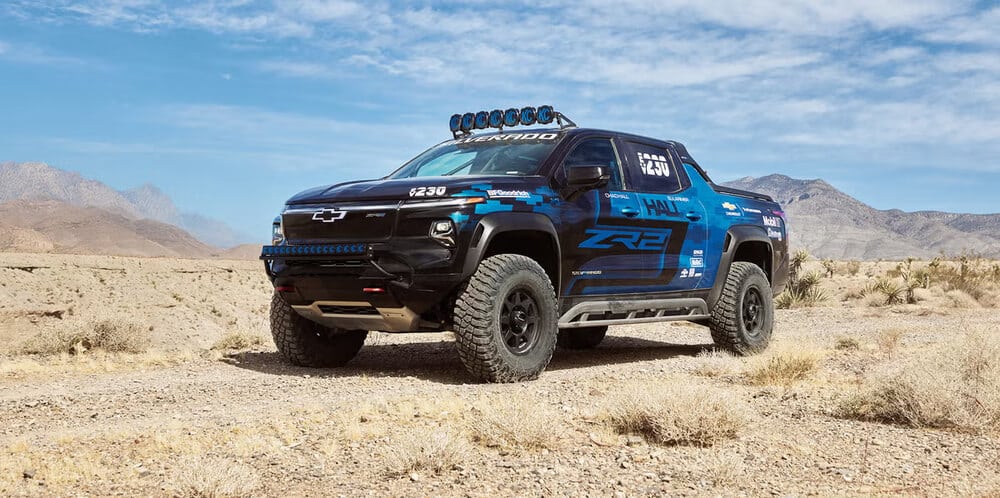 "Una camioneta Chevrolet Silverado EV ZR2 se encuentra estacionada en un paisaje desértico con montañas al fondo. La camioneta es negra con detalles en azul, tiene neumáticos todoterreno grandes, una barra de luces en el techo y varios adhesivos de patrocinadores. El cielo está despejado con algunas nubes, y el terreno es seco y rocoso.