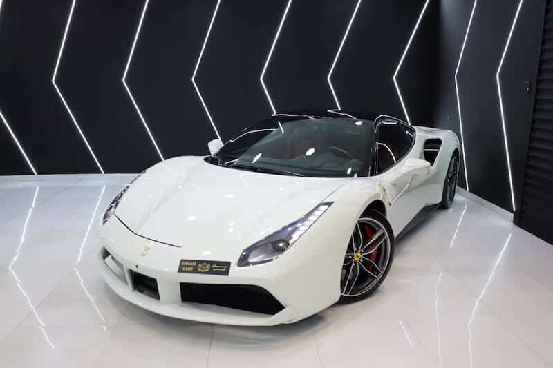 "Imagen de un Ferrari 488 Spider blanco en un showroom moderno con diseño de luces LED en forma de zigzag en las paredes. Este coche de lujo es representativo de los vehículos de alta gama que pueden ser importados de Dubái a España, ideales para compradores interesados en calidad y exclusividad.