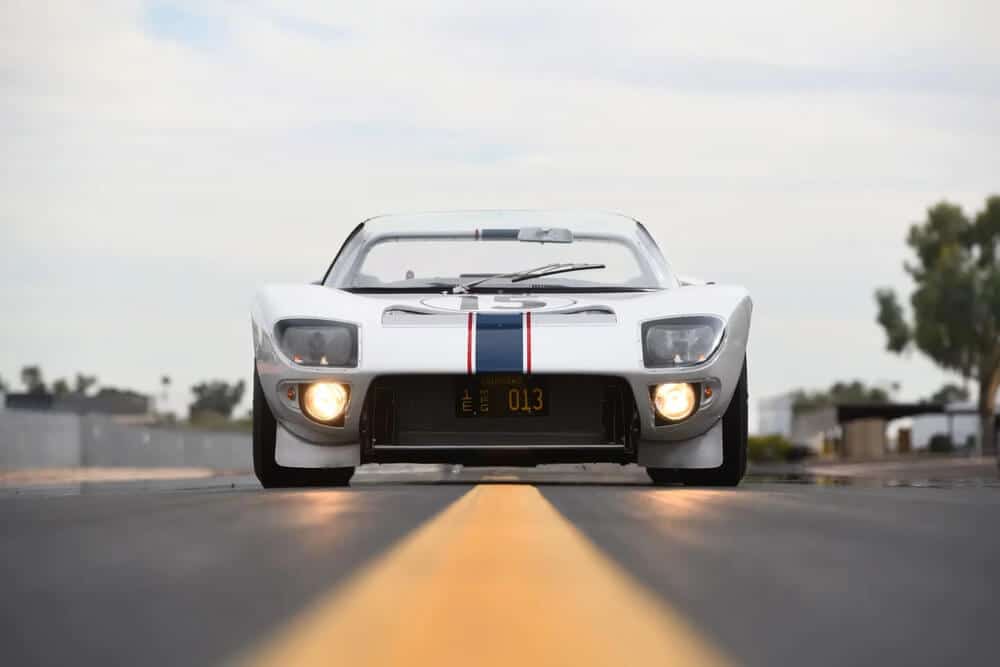 "Ford GT40 descapotable blanco visto de frente, con franjas azul y roja en el centro del capó, el número 5 en la parte superior, y matrícula con el número '013'. El coche está ubicado en una carretera con línea amarilla central, rodeado de árboles y edificios al fondo. Modelo icónico en subasta.