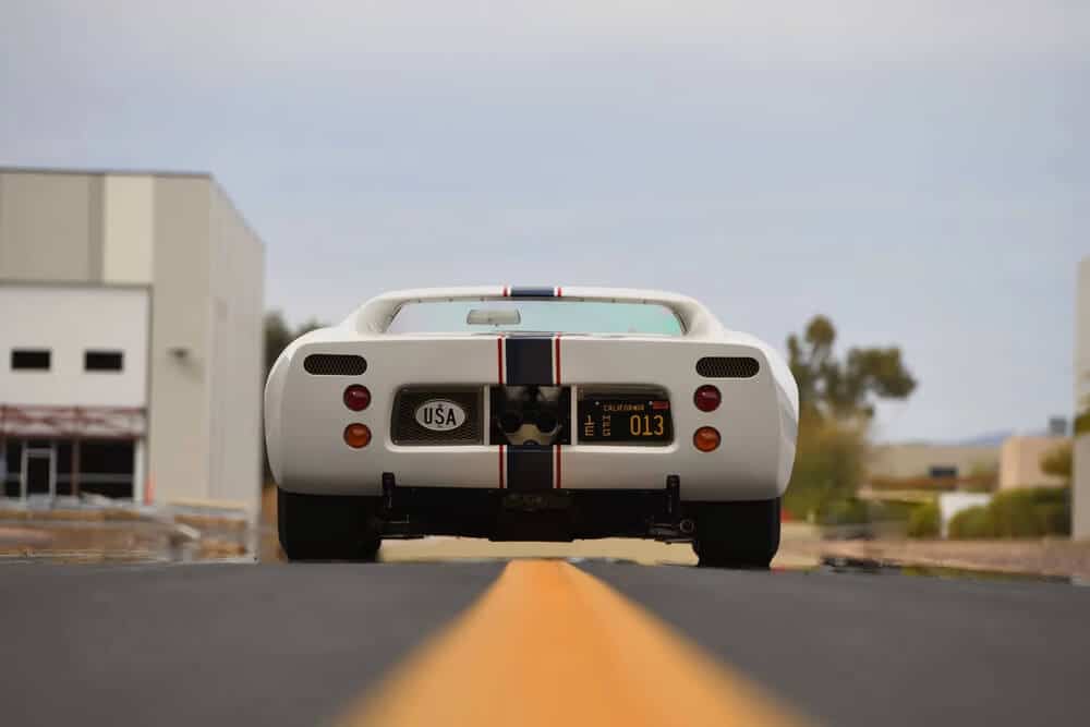 "Vista trasera del Ford GT40 descapotable blanco, con franjas verticales azul y roja recorriendo el centro del coche. La matrícula muestra 'California 013' y lleva una etiqueta que dice 'USA'. Este emblemático modelo está estacionado sobre una carretera asfaltada con línea amarilla en el centro, rodeado de árboles y edificios en el fondo. Este icónico coche deportivo se encuentra actualmente en subasta.