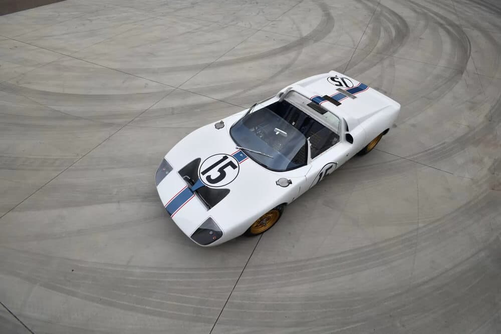 Ford GT40 descapotable blanco con franjas azul y roja atravesando el centro del coche, con el número 15 en círculos negros visibles en los laterales y el capó. Equipado con llantas doradas y diseño deportivo, el coche se muestra en una carretera rodeada de árboles y paisajes al fondo. Actualmente, este modelo emblemático está siendo subastado.