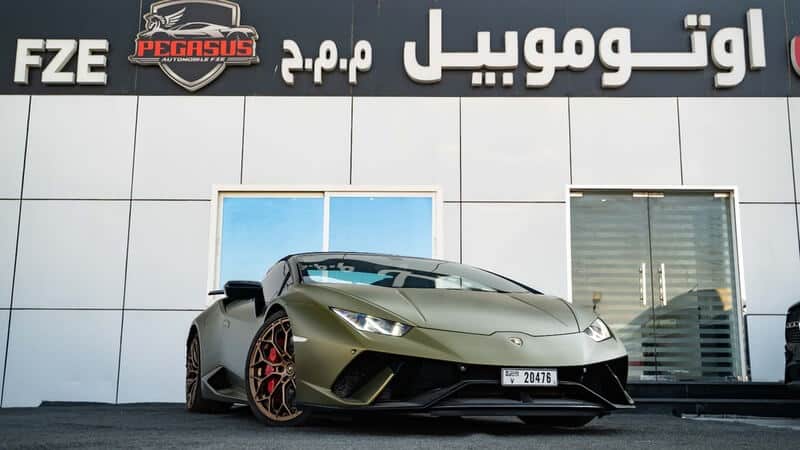 "Imagen de un Lamborghini Huracán EVO Spyder en color verde mate, estacionado frente a un concesionario llamado Pegasus Automobile FZE en Dubái. El coche tiene un diseño deportivo y elegante, destacando sus líneas aerodinámicas y llantas negras. Esta imagen está relacionada con el keyword SEO 'importar coches de Dubái a España', mostrando un vehículo de lujo que podría interesar a compradores españoles.