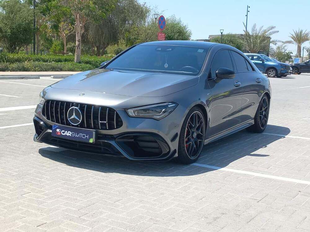 Imagen de un coche Mercedes-Benz AMG CLA 45 estacionado en un aparcamiento en Dubái. El coche es de color gris metálico, con un diseño deportivo que incluye llantas negras y detalles en rojo. Esta imagen es relevante para el keyword SEO 'importar coches de Dubái a España', ya que muestra un vehículo de alta gama que podría interesar a importadores y compradores en España.