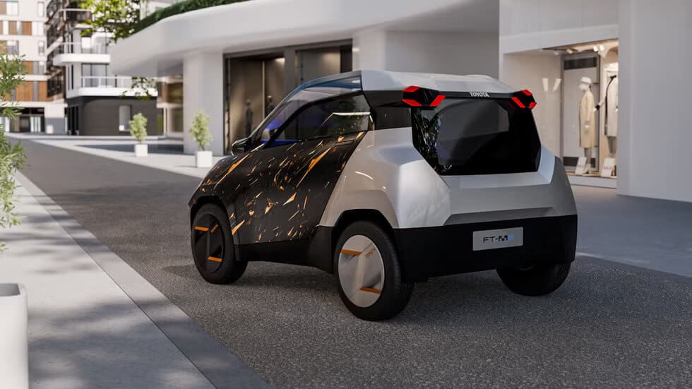 Imagen de un automóvil compacto y futurista, el Toyota FT-M. El coche tiene un diseño moderno con líneas angulares y una combinación de colores en negro, naranja y plateado. Las luces traseras tienen forma de flecha y las ruedas destacan con detalles en naranja. El coche está estacionado en una calle de una ciudad moderna, rodeado de edificios y tiendas contemporáneos.