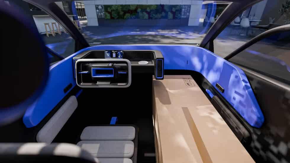 Imagen del interior del Toyota FT-M. El diseño es futurista y minimalista, con un tablero de instrumentos digital y centrado, acompañado por una pantalla táctil rectangular. Los asientos son de color gris claro y parecen ser ergonómicos y cómodos. Hay una superficie plana amplia que podría servir para almacenamiento o como espacio de trabajo. La iluminación azul ambiental del interior aporta un toque tecnológico y moderno, mientras que a través de las ventanas se observa un paisaje urbano con vegetación.