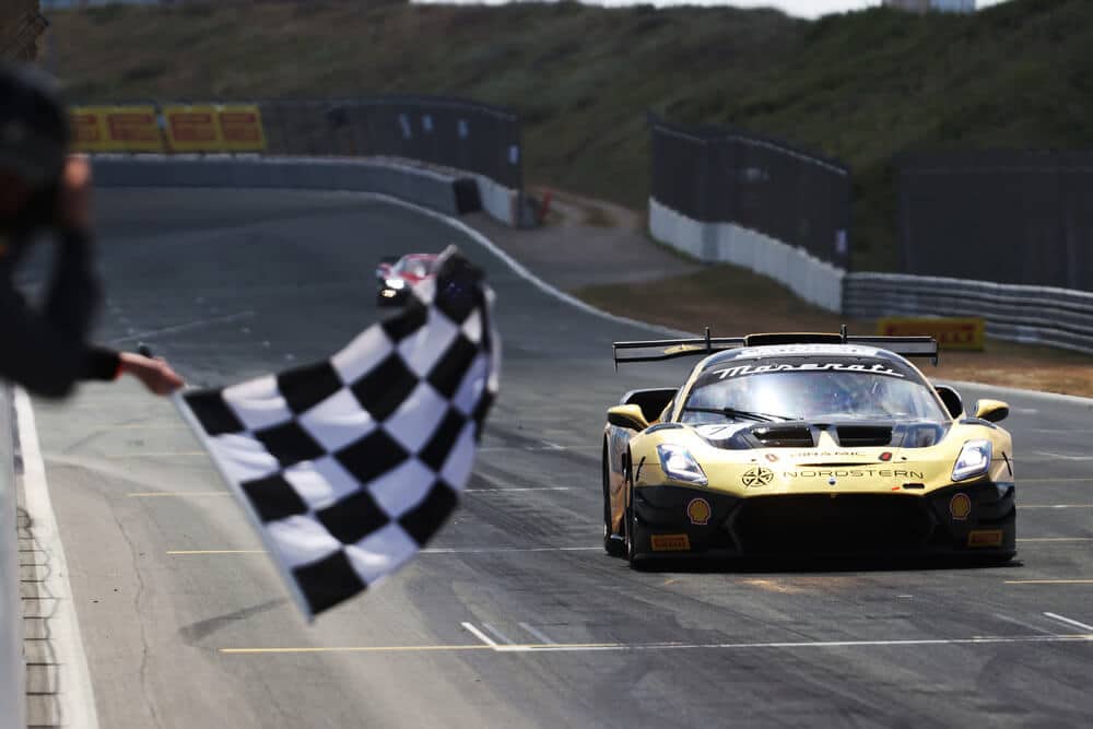 Un coche de carreras Maserati, con un diseño aerodinámico y agresivo, avanza a toda velocidad por la pista. La carrocería es negra con detalles en rojo y azul, y exhibe los logotipos de Maserati, apm Monaco, Shell y Pirelli. Las ruedas Pirelli están en contacto con el asfalto mientras el vehículo toma una curva cerrada, transmitiendo la emoción y la intensidad de la competición.