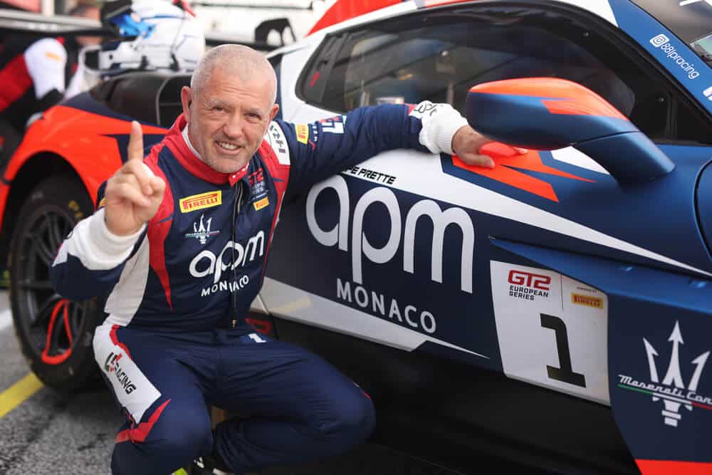 "La imagen muestra dos coches de carreras Maserati en una pista de competición. El coche en primer plano es dorado con detalles en negro, y luce los logotipos de Shell, Pirelli, Dinamic y Nordstern en la parte delantera. Su diseño aerodinámico y su gran alerón trasero resaltan su enfoque en la velocidad y estabilidad. Detrás, otro Maserati negro con detalles en rojo y azul exhibe los logotipos de Shell, Pirelli y apm Monaco. Ambos vehículos están en plena acción, inclinándose en una curva cerrada, transmitiendo la intensidad de la competencia y la emoción del automovilismo.