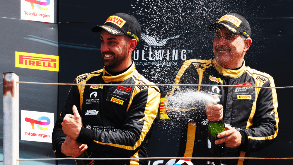 Dos pilotos de carreras celebran en el podio. Uno de ellos, vestido con un mono de competición con logotipos de Pirelli, TotalEnergies y GT2 European Series, levanta el pulgar en señal de victoria. El otro piloto sostiene una botella de champán y la rocía en el aire, capturando la emoción del momento. En el fondo, se aprecia el logo de 'Gullwing Racing Insurance', reforzando el ambiente de celebración en el mundo del automovilismo.