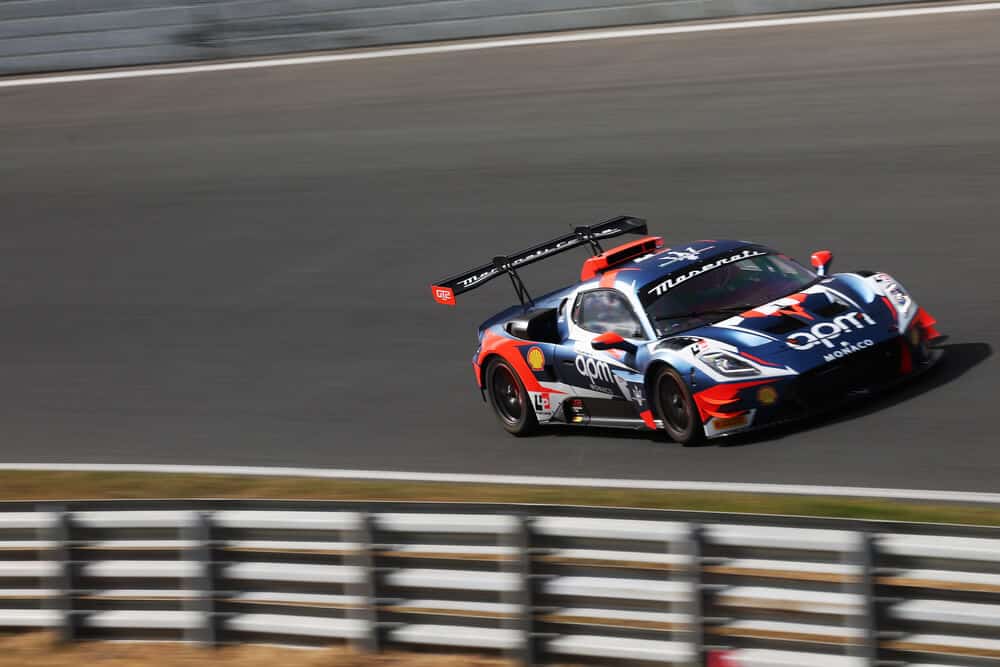 Un único coche de carreras Maserati en acción sobre la pista. Se trata de un vehículo con un diseño aerodinámico, de color negro con detalles en rojo y azul, que lleva los logotipos de Maserati, apm Monaco, Shell y Pirelli en su carrocería. Sus neumáticos Pirelli están en contacto con el asfalto mientras toma una curva, transmitiendo la emoción y la velocidad propias de la competición.