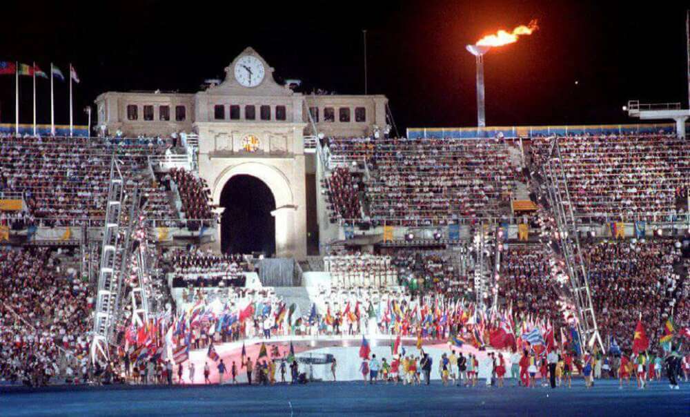 Imagen de la ceremonia de apertura de los Juegos Olímpicos de Barcelona 1992. El estadio está lleno de espectadores, con una estructura arquitectónica prominente que incluye un arco y un reloj. En la parte superior derecha, se observa la llama olímpica encendida. En la parte inferior, una multitud de personas porta banderas de diferentes países, representando la diversidad de naciones participantes.