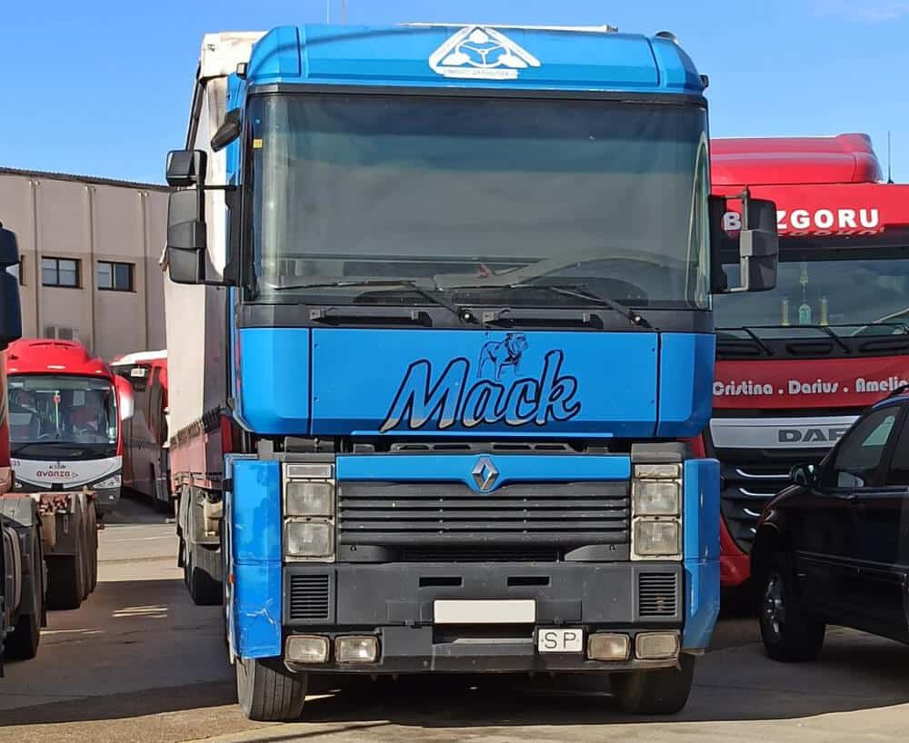 Camión Renault azul con la palabra 'Mack' en letras grandes y estilizadas en la parte frontal, acompañado por un dibujo de un bulldog sobre el texto. Se encuentra estacionado en un área con otros vehículos grandes, incluidos autobuses y camiones. En el fondo, se distingue un camión rojo con la palabra 'BIZGORU' en la parte superior del parabrisas y un autobús rojo y blanco con 'avanza' escrito en la parte frontal.
