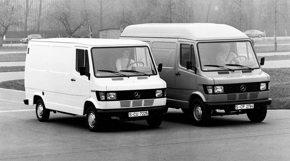 La imagen muestra dos furgonetas Mercedes-Benz estacionadas una al lado de la otra. La furgoneta de la izquierda es de color blanco y tiene la matrícula 'S-CU 7225'. La furgoneta de la derecha es de color gris y tiene la matrícula 'S-CP 2784'. Ambas furgonetas son modelos Mercedes-Benz T1, también conocidos como Bremer Transporter. La furgoneta blanca parece ser una versión estándar, mientras que la furgoneta gris tiene un techo elevado, lo que indica que podría ser una versión de mayor capacidad.