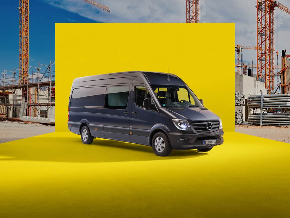 La imagen muestra una furgoneta Mercedes-Benz Sprinter de color gris oscuro, estacionada sobre una superficie amarilla brillante. Al fondo, se observa un sitio de construcción con grúas y estructuras en proceso de edificación. La matrícula de la furgoneta es 'S MB 6211'. Por su diseño moderno, líneas aerodinámicas y características visibles, parece ser un modelo de los años 2018 a 2020.