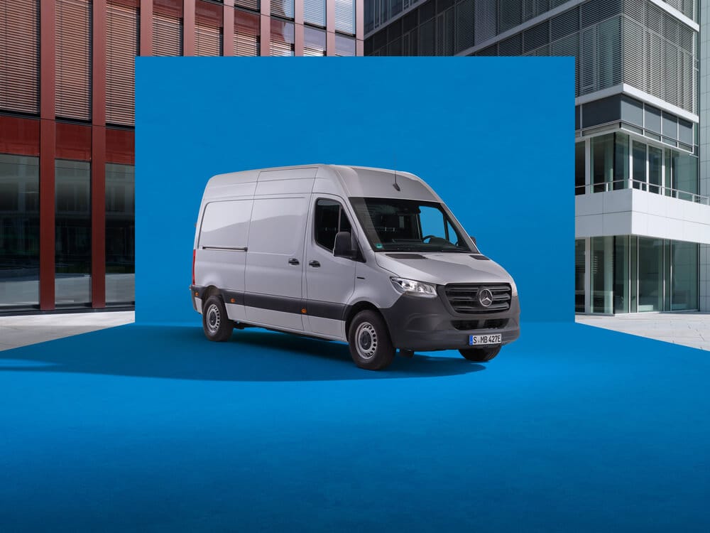 La imagen muestra una furgoneta Mercedes-Benz Sprinter de color gris, estacionada sobre una superficie azul brillante. El fondo azul contrasta con los edificios modernos de vidrio y metal que la rodean. La matrícula de la furgoneta es 'S-MB 427E'. Por su diseño y las características visibles, parece ser un modelo de los años 2022 a 2024.