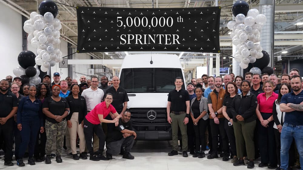 Un grupo de personas posa frente a una camioneta Mercedes-Benz Sprinter blanca en una fábrica. Sobre la camioneta hay un cartel negro con texto blanco que dice '5,000,000th SPRINTER'. A ambos lados del cartel hay arreglos de globos blancos y negros. La imagen celebra la producción de la camioneta número cinco millones del modelo Sprinter.