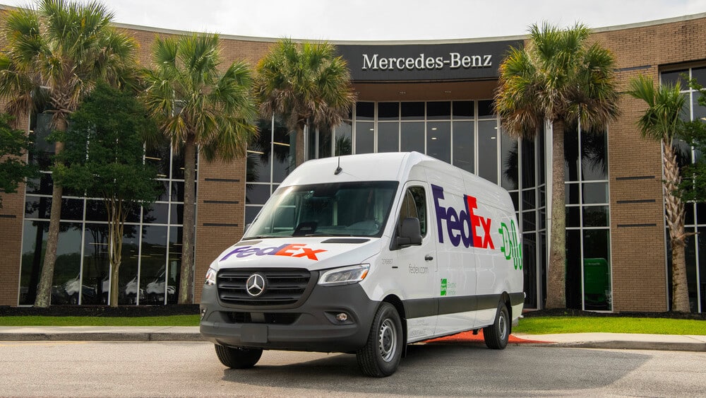 "La imagen muestra una furgoneta Mercedes-Benz Sprinter con el logotipo de FedEx estacionada frente a un moderno edificio de Mercedes-Benz. La furgoneta, diseñada para entregas, tiene las palabras 'Electric Vehicle' y 'fedex.com' en el costado. Por su estilo aerodinámico y detalles visibles, parece ser un modelo del año 2023. El fondo del edificio de Mercedes-Benz, adornado con palmeras, refuerza el contexto de innovación y tecnología.