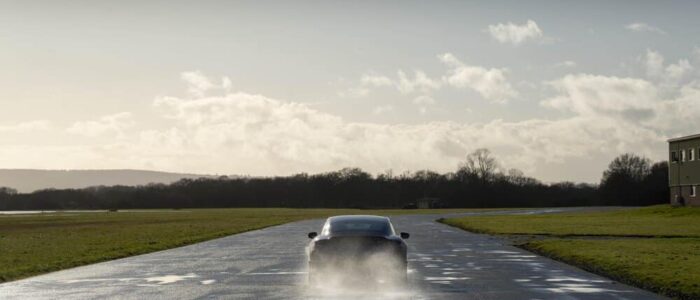 Un Porsche Taycan azul está circulando sobre una carretera mojada, levantando agua a su paso. A los lados del camino hay campos de césped, y a la derecha se distingue un edificio. El cielo está parcialmente nublado, con algunos rayos de sol atravesando las nubes, creando una atmósfera brillante y clara.