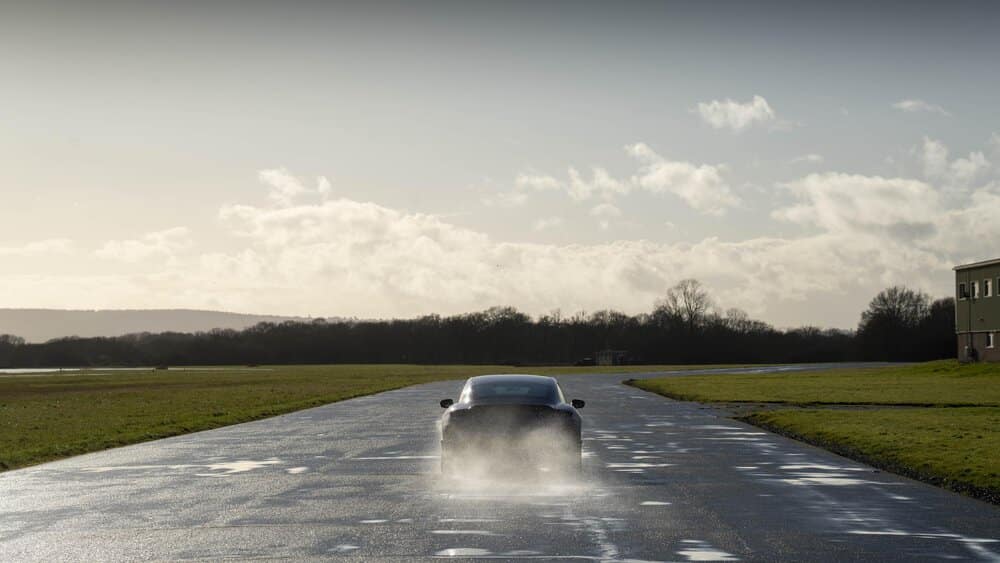 Un Porsche Taycan azul está circulando sobre una carretera mojada, levantando agua a su paso. A los lados del camino hay campos de césped, y a la derecha se distingue un edificio. El cielo está parcialmente nublado, con algunos rayos de sol atravesando las nubes, creando una atmósfera brillante y clara.