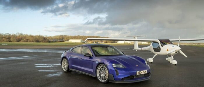 Un Porsche Taycan azul está aparcado sobre una pista de aterrizaje, con un planeador en primer plano y otro en el fondo. La cola del planeador en primer plano muestra la matrícula 'G-KDKD' y una bandera británica. La imagen evoca el estilo de pruebas y comparaciones típicas de Top Gear, donde la velocidad y la tecnología se encuentran en entornos inesperados.