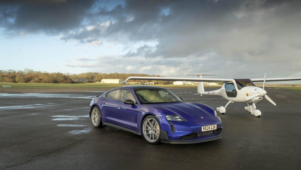 Un Porsche Taycan azul está aparcado sobre una pista de aterrizaje, con un planeador en primer plano y otro en el fondo. La cola del planeador en primer plano muestra la matrícula 'G-KDKD' y una bandera británica. La imagen evoca el estilo de pruebas y comparaciones típicas de Top Gear, donde la velocidad y la tecnología se encuentran en entornos inesperados.