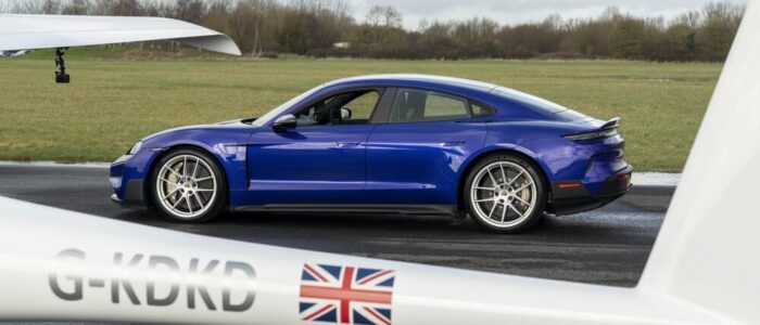 Un Porsche Taycan azul está aparcado sobre el asfalto de una pista de aterrizaje. En primer plano, la cola y el ala de un planeador con matrícula 'G-KDKD' y una bandera británica en la cola destacan en la escena. Al fondo, se ven áreas de césped y árboles, sugiriendo un entorno de aeródromo.