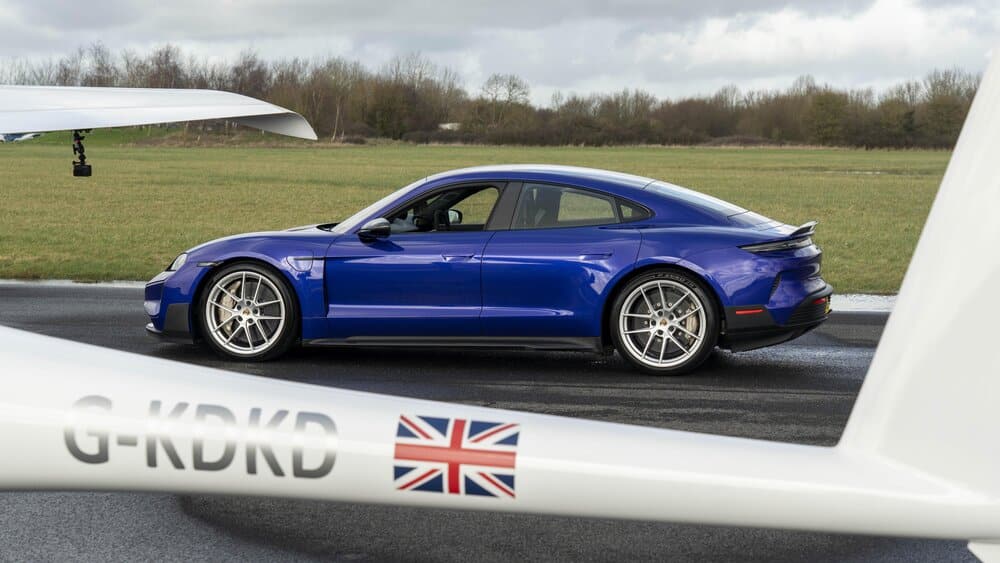 Un Porsche Taycan azul está aparcado sobre el asfalto de una pista de aterrizaje. En primer plano, la cola y el ala de un planeador con matrícula 'G-KDKD' y una bandera británica en la cola destacan en la escena. Al fondo, se ven áreas de césped y árboles, sugiriendo un entorno de aeródromo.