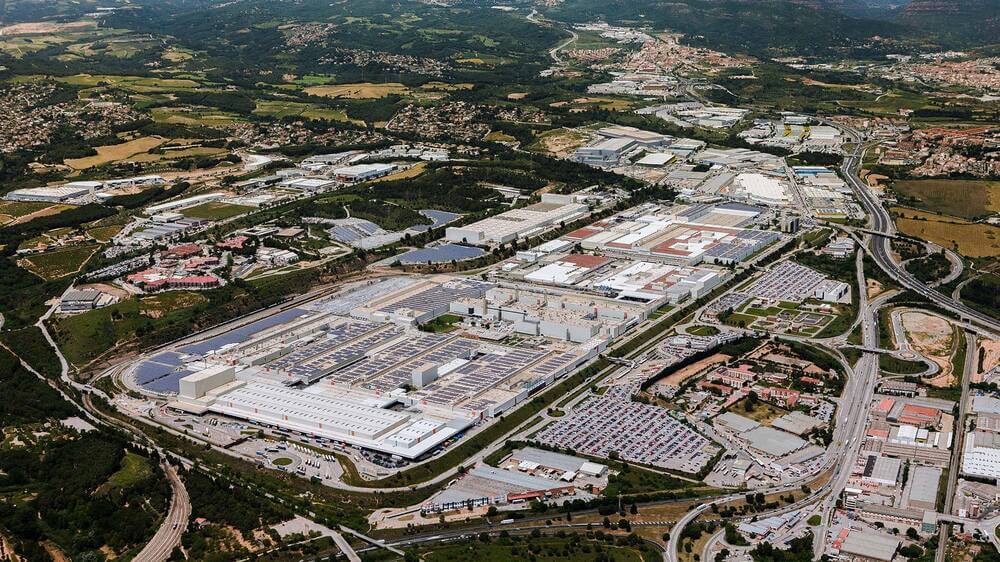 Vista aérea de la planta de producción de Seat en Martorell, España. La fábrica ocupa una gran extensión de terreno y está rodeada por carreteras y áreas industriales. Se observan numerosos edificios y estructuras que forman parte del complejo, así como zonas de estacionamiento llenas de vehículos. En el fondo, hay áreas verdes y residenciales, mostrando la integración de la planta en su entorno.